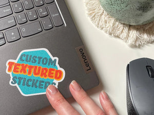 A custom textured sticker with the text 'CUSTOM TEXTURED STICKER' placed on a laptop next to a mouse, on a desk.