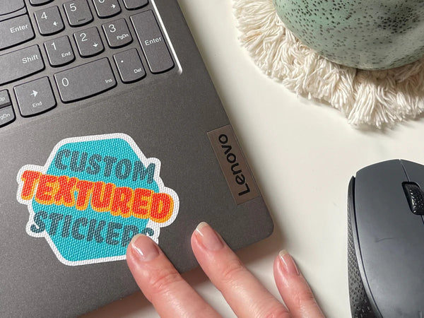 A custom textured sticker with the text 'CUSTOM TEXTURED STICKER' placed on a laptop next to a mouse, on a desk.